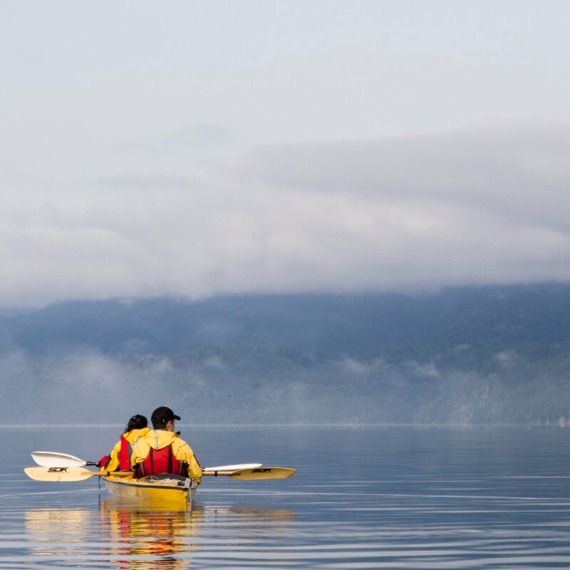 Two people in a yellow kayak paddle on a calm lake with foggy, tree-covered mountains in the distance. Luxury Argentina tours.