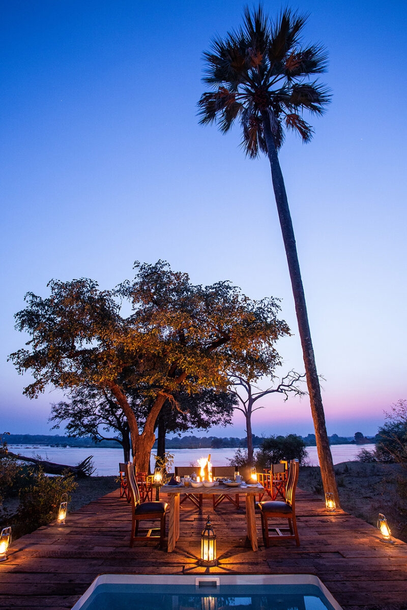 A wooden deck set for a dinner, lit by lanterns under a tall palm tree with a river and sunset in the background.