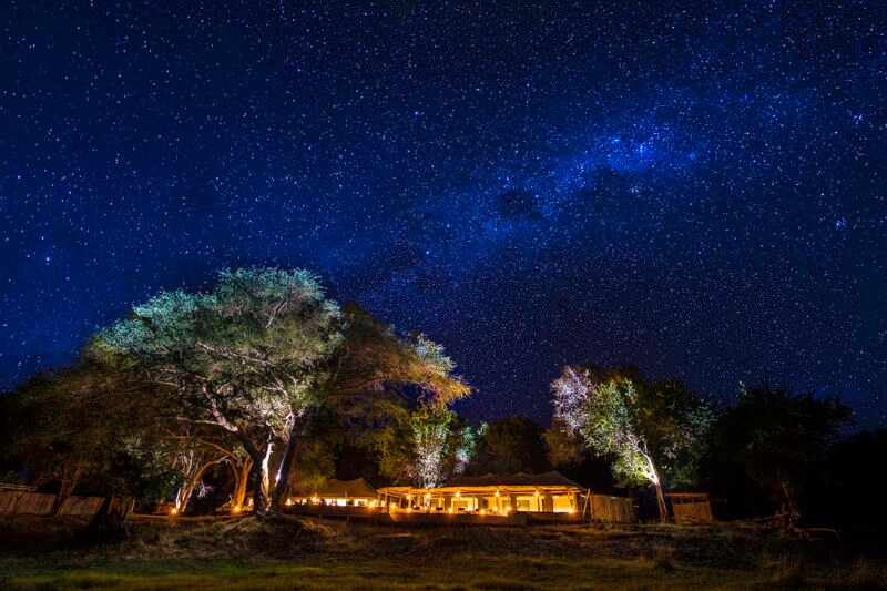 An illuminated luxury safari camp, visible under a spectacular dark night sky full of bright stars and the Milky Way.