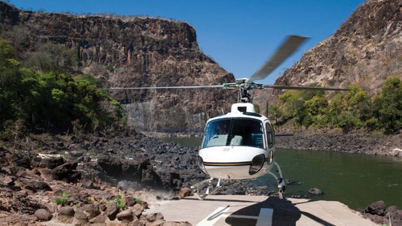 A white helicopter lifting off a landing pad on a rocky bank next to a river and steep, rocky cliffs.