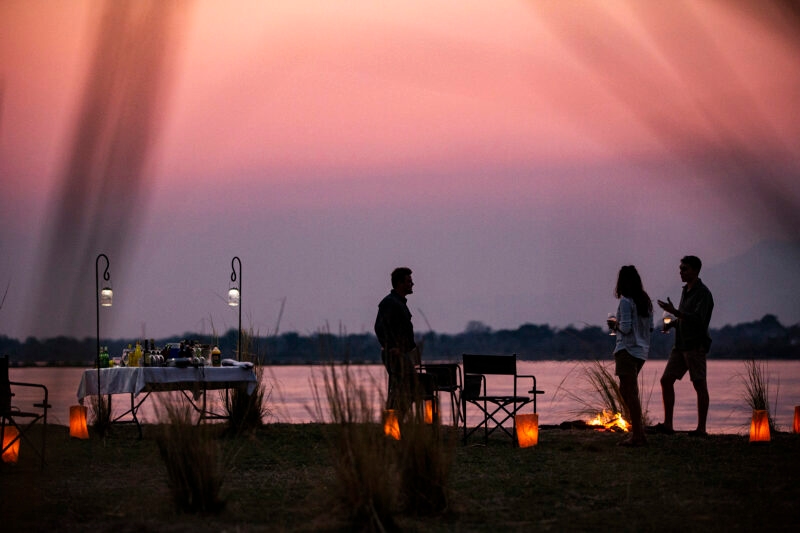 Silhouettes of three people standing by a river at sunset, with a bar table and lanterns lit on the grassy bank.