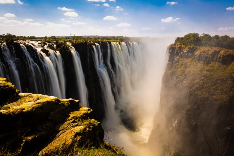 Victoria Falls, with a massive amount of water plunging into a deep, misty gorge under a blue sky.