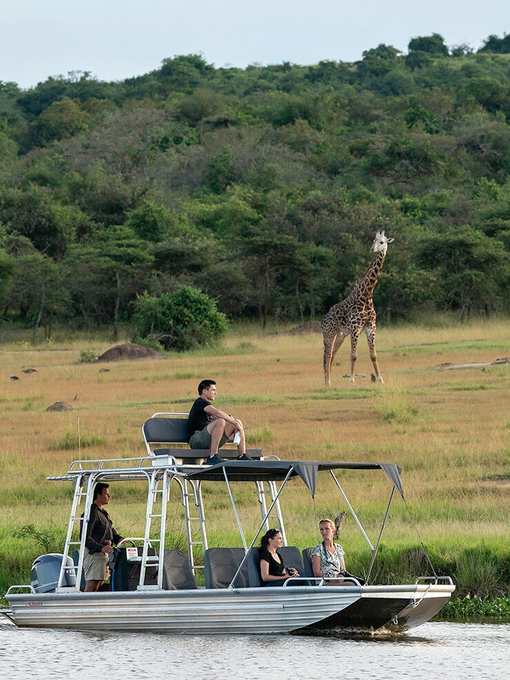 Tourists on a covered boat watching a giraffe on the grassy riverbank in the distance. Book luxury Rwanda safaris today.