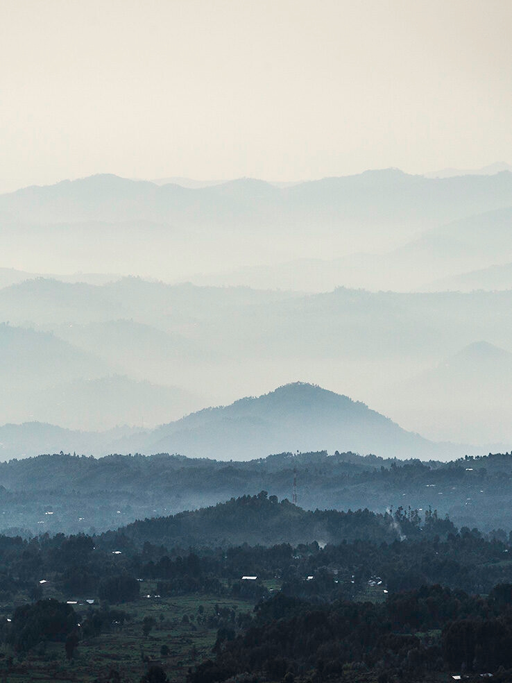 Wide view of tiered rolling hills and valleys covered in layers of blue and white mist. Enjoy the landscapes on luxury Rwanda vacations.