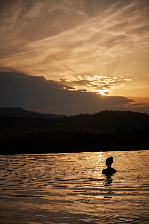 Luxury Italy Tours - The silhouette of the head a shoulders of a woman with long hair piled on top of her head in a bun style, sitting in calm water overlooking hills and mountains during a cloudy, golden sunset