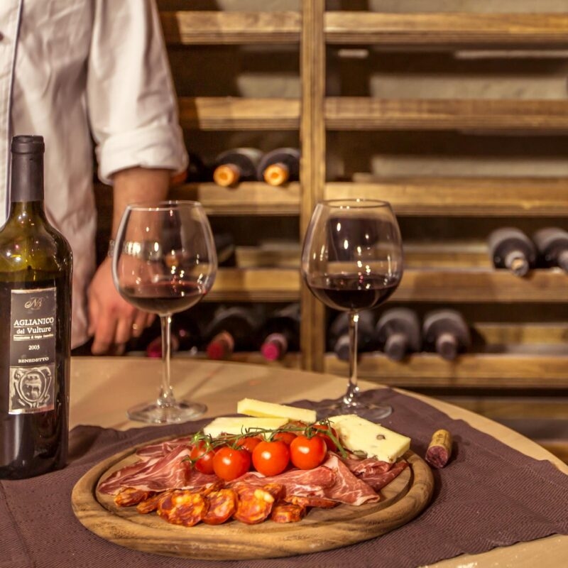 Luxury Italy Tours - A wooden round plate on a table laden with fresh small tomatoes, cured meats, and cheese. There are also two wine glasses with a small amount of red wine, and a green wine bottle with the cork laid next tot he plate. In the background, there are rows of wine bottles laying on wooden shelves in a cellar, and we see the arm of a person wearing a chef's white jacket