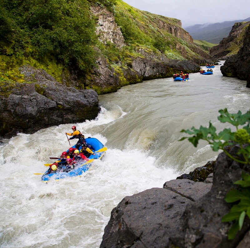 Adventurers in blue rafts paddle through white water rapids in a deep canyon surrounded by green cliffs.