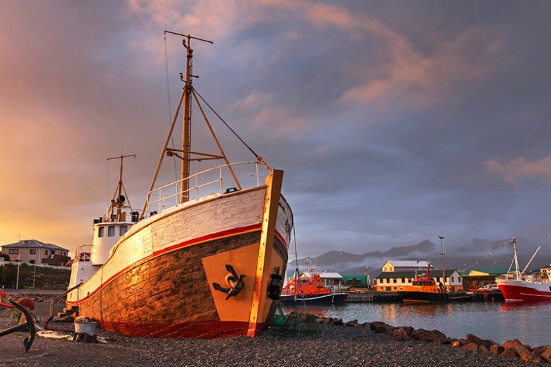 A large white and brown boat on a rocky shore in a harbor under a sunset sky.