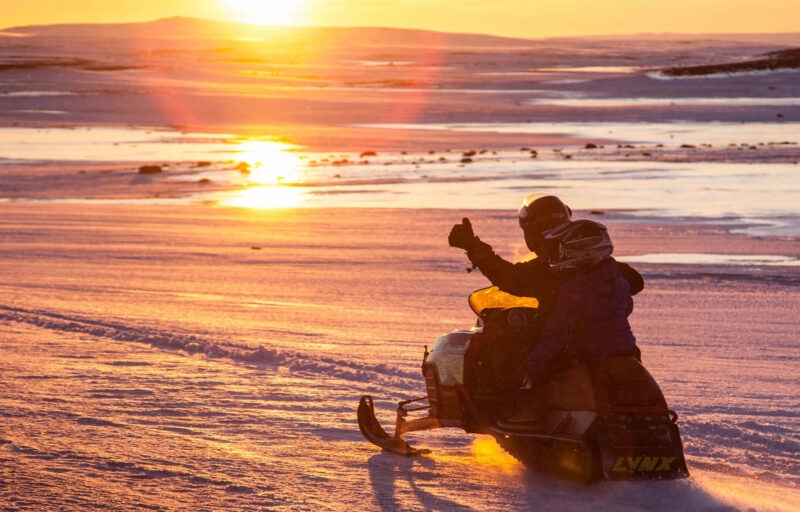 Silhouette of two people on a snowmobile at sunset, with one person giving a thumbs up.