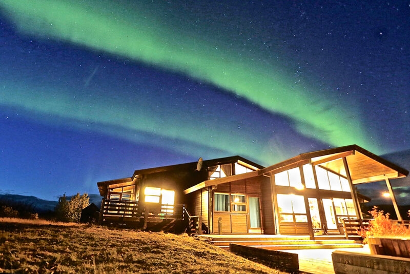 A modern wooden cabin glows at night under the bright green Aurora Borealis, an ideal setting for luxury Iceland holidays.