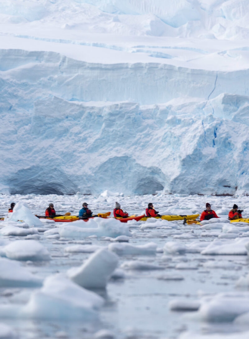 Several people in yellow kayaks are paddling near a massive blue and white glacier wall with floating ice chunks.