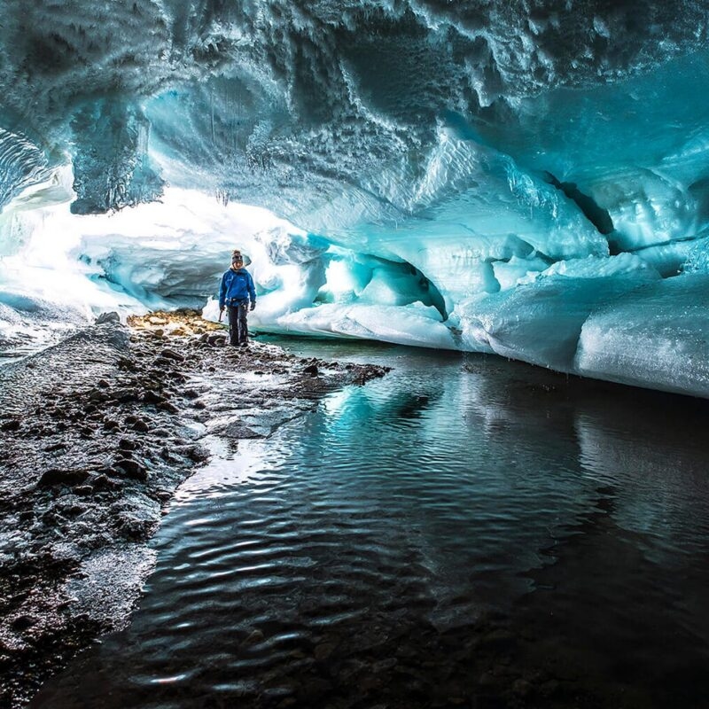 A person stands inside a glowing blue glacier ice cave next to a stream of water on the dark floor.