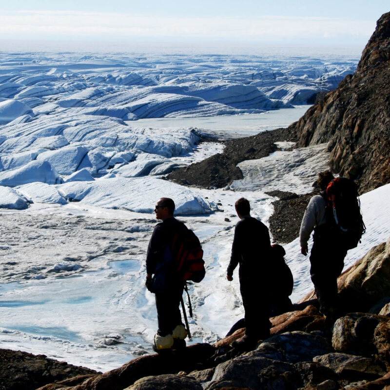 Three hikers stand on a rocky hill overlooking a vast, white and blue glacier landscape.