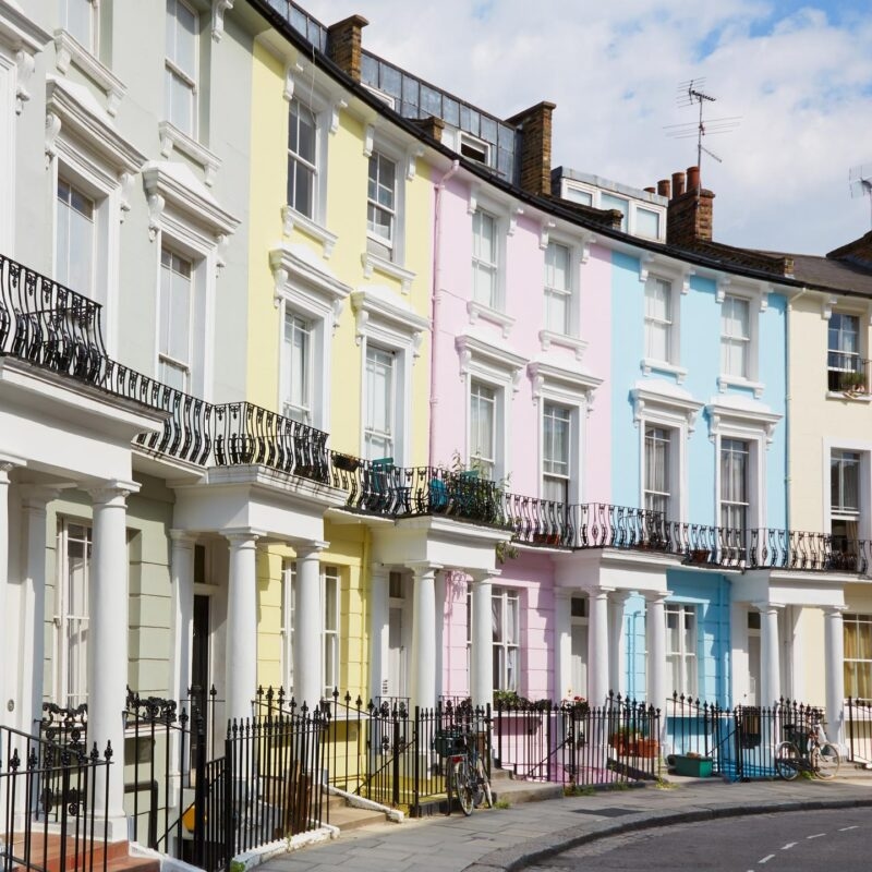 Row of pastel-colored terraced houses in London with white columns, black railings, and small balconies on a sunny day.