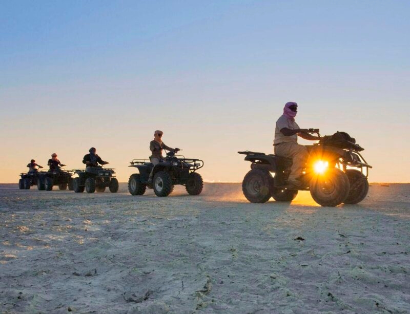 Five people riding ATVs in a row across a sandy desert at sunset with a clear sky background.
