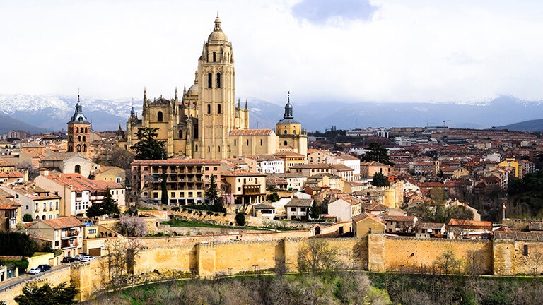 Panoramic view of the city of Segovia with its cathedral, a stone wall, and snowy mountains. Book luxury Spain trips.