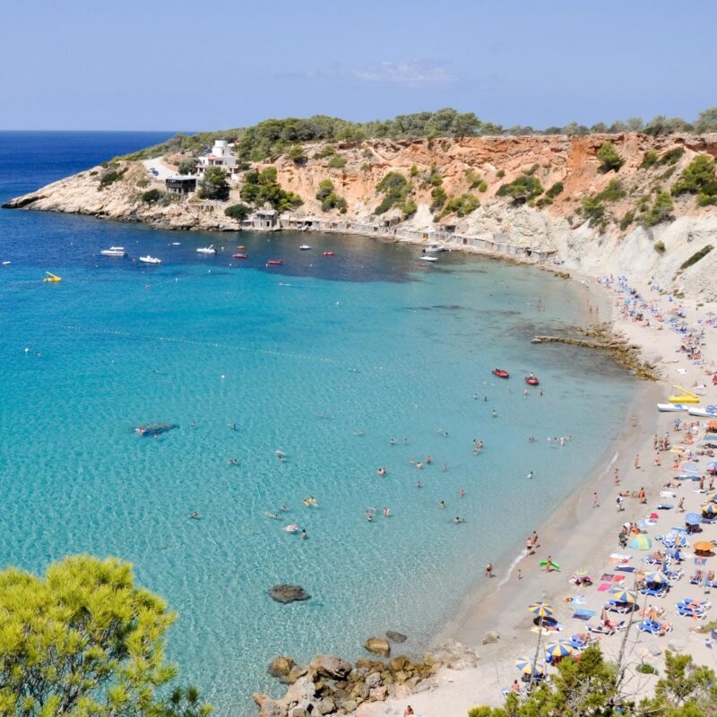 Aerial view of a crowded beach on a turquoise bay backed by a red cliff. A perfect location for luxury Spain trips.