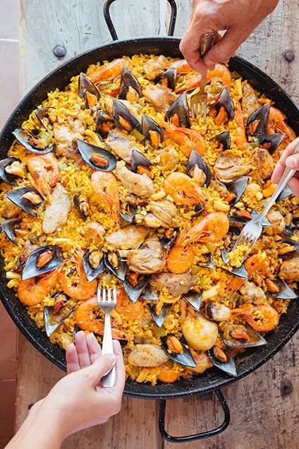 Overhead view of a large pan of seafood paella with several hands holding forks, representing luxury Spain tours.
