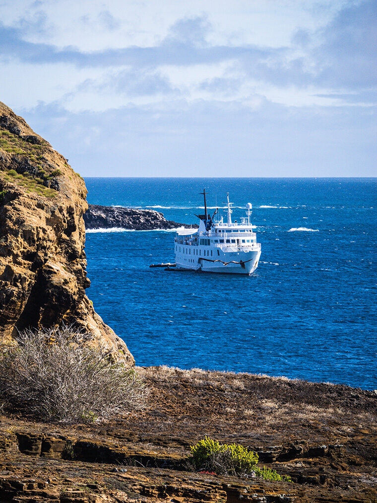 A white cruise ship sailing on the blue ocean near a rocky coast under a blue sky with some white clouds. luxury Galapagos cruises.