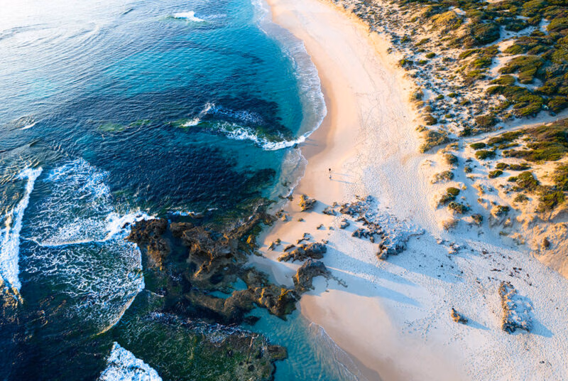 Aerial view of a rugged, curved coastline with white sand, turquoise water, and breaking waves, a beautiful stop on luxury Australia trips.