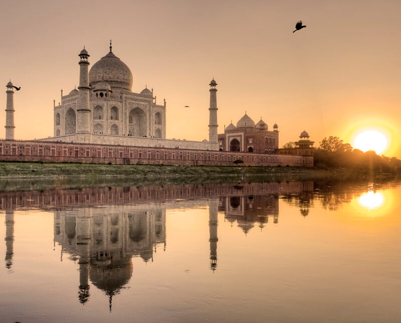 The Taj Mahal is reflected in the water at sunset, with a vibrant orange sun on the horizon, a view from a luxury India tours.