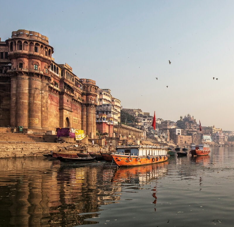 Traditional stone ghats and buildings line the Ganges River, with several boats docked in the foreground in Varanasi on luxury India tours.