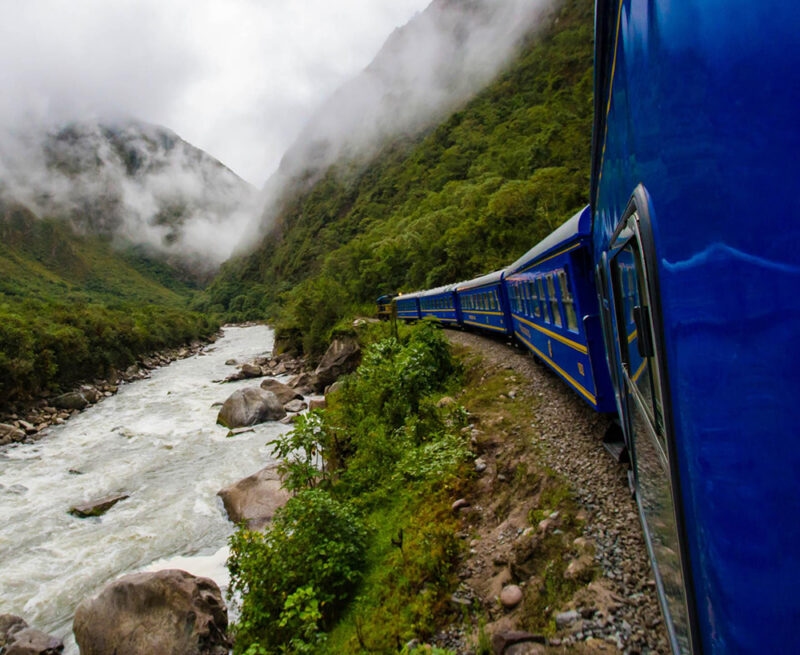 A blue train rounding a curve by a river on luxury Peru train tours.