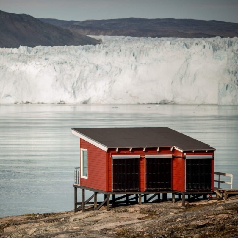 A red wooden cabin on stilts near water and a giant glacier during luxury Polar vacations.