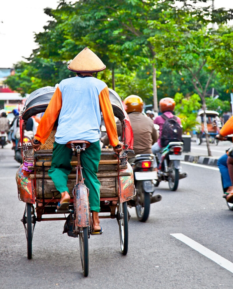 A man in a conical hat and bright shirt rides a becak (cycle rickshaw) down a street. Traffic and trees are in the background, offering luxury Indonesia tours.