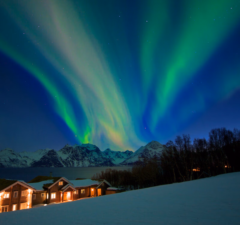 Brilliant green and blue Northern Lights above snow-covered mountains and illuminated wooden cabins, a highlight of luxury Norway holidays.