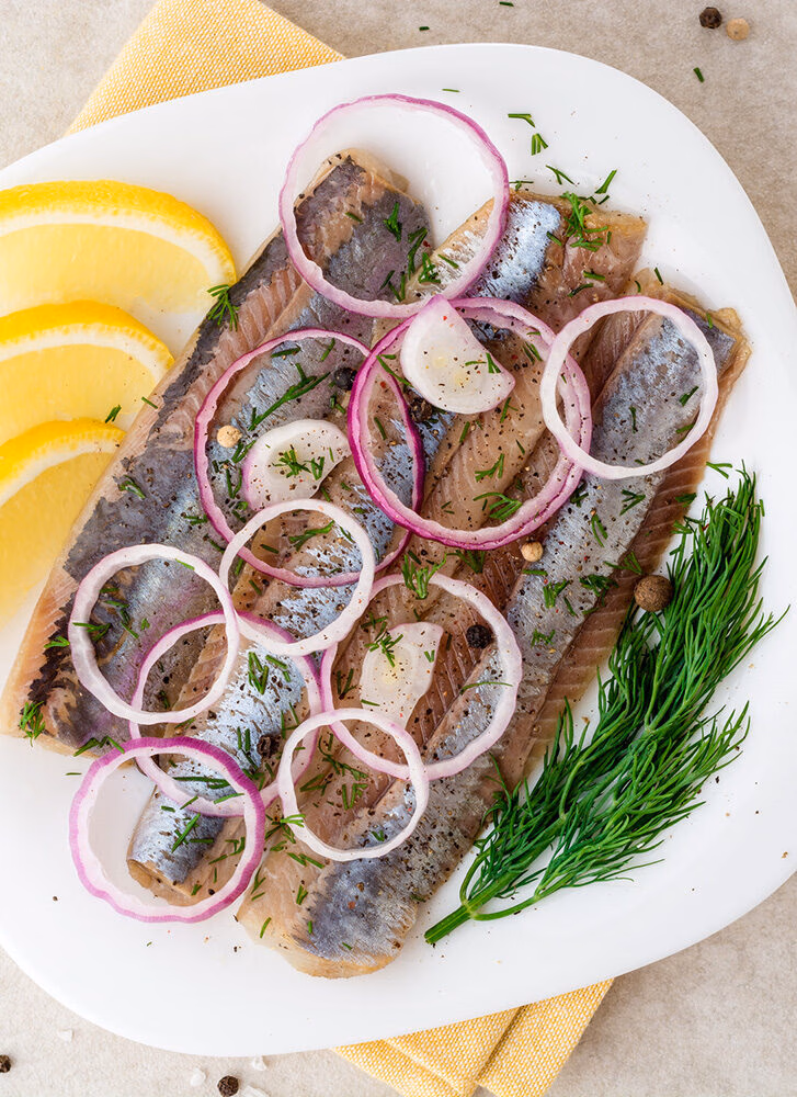 Pickled herring fillets on a white plate, garnished with red onion rings, dill, and lemon wedges, typical for luxury Norway trips.