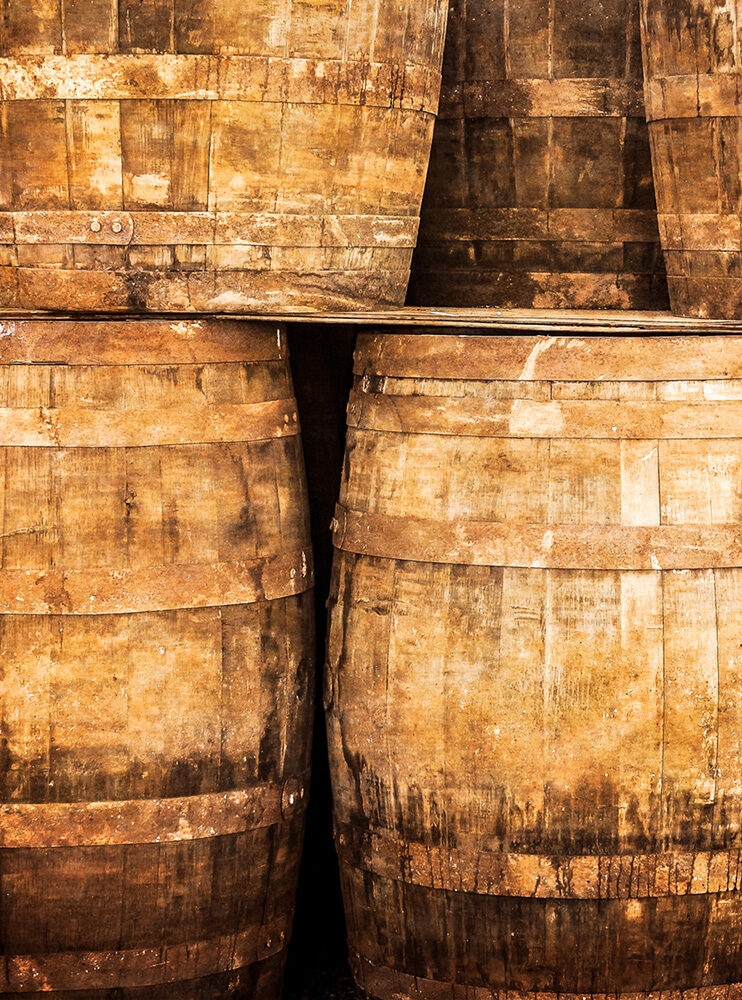 A close-up image of rough, aged wooden barrels stacked tightly together in a warm, brown-orange tone. Explore this aspect of luxury Scotland vacations.