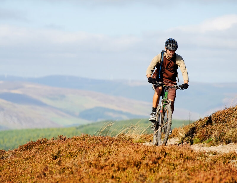 A man rides a mountain bike up a dusty, scrub-covered hill, with a vast, blue-skied mountain landscape behind. Perfect for luxury Scotland trips.