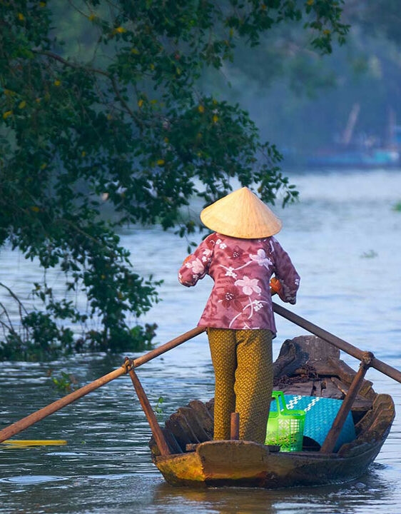Luxury Vietnam Tours - A person in traditional Vietnamese clothing standing on a shallow boat with two long oars, paddling through calm water