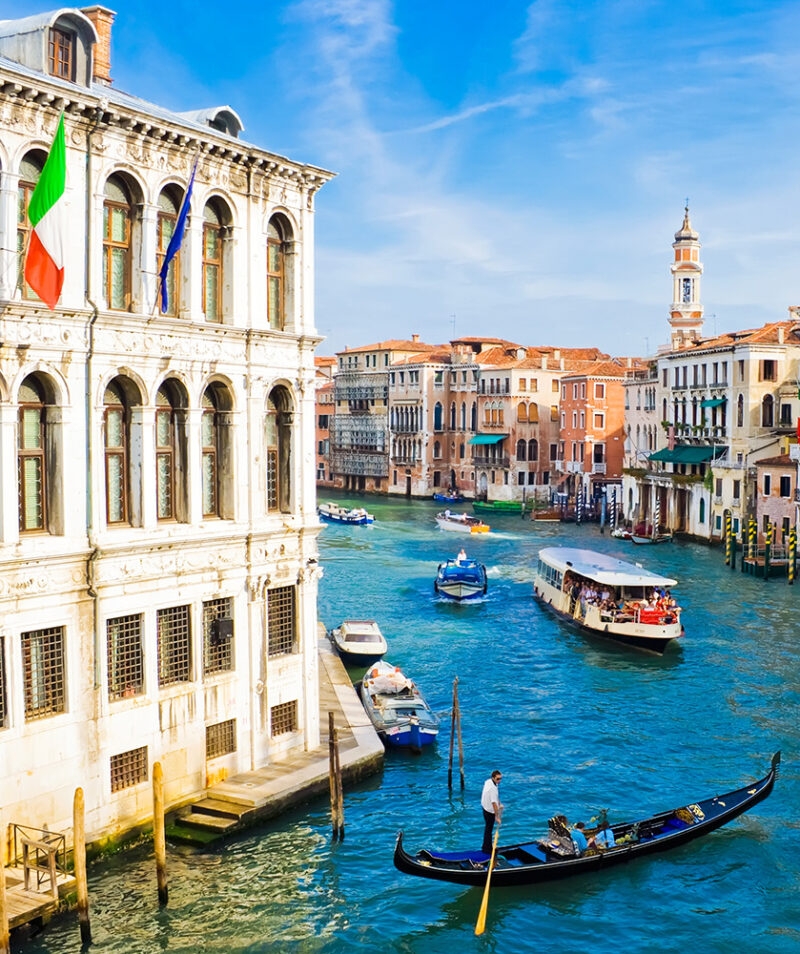 A man in a gondola on the bright blue Grand Canal in Venice, Italy, framed by a white historic palace and colorful buildings.