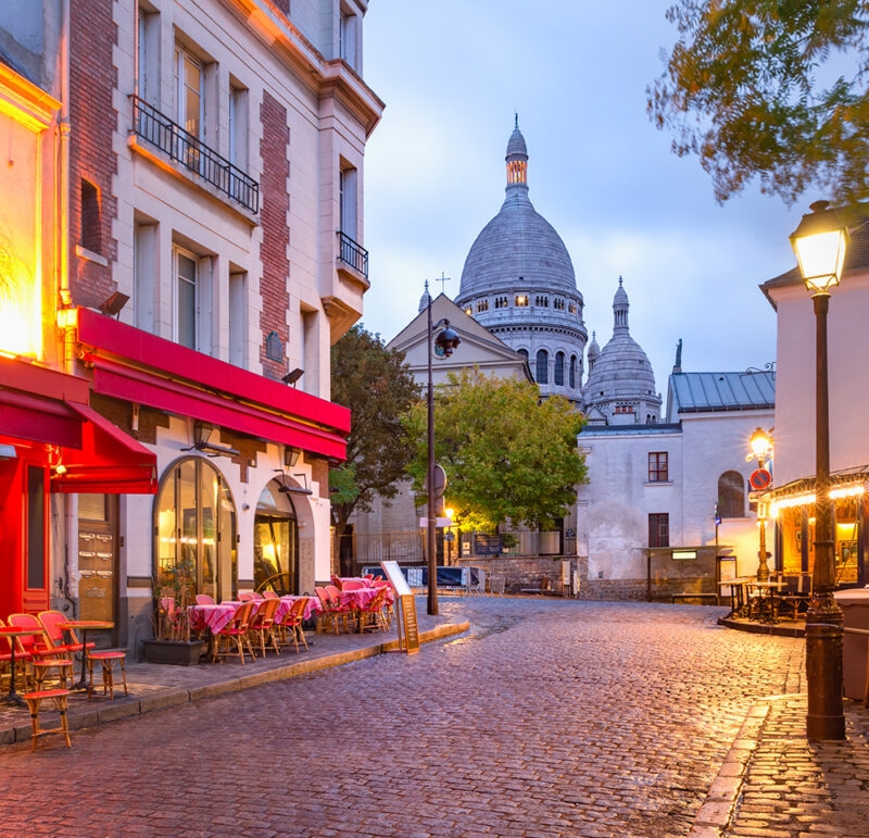 A quiet, cobblestone street in Montmartre, Paris, featuring illuminated café terraces and the domes of the Sacré-Cœur Basilica at dusk.