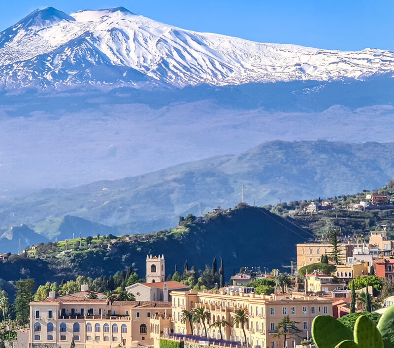 The snow-capped Mount Etna volcano dominates the background above the colorful townscape of Taormina, Sicily, Italy.