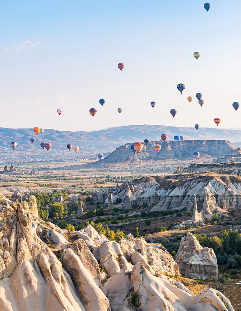 Dozens of colorful hot air balloons float over the dramatic, rocky landscape of Cappadocia, Turkey, featuring unique "fairy chimney" formations.