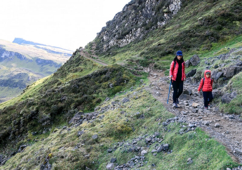 A woman and a small child in red jackets hiking on a steep, rocky mountain trail under a bright sky.