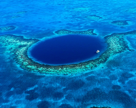 An aerial shot of a large, circular dark blue underwater sinkhole surrounded by turquoise coral reef and a small white boat.