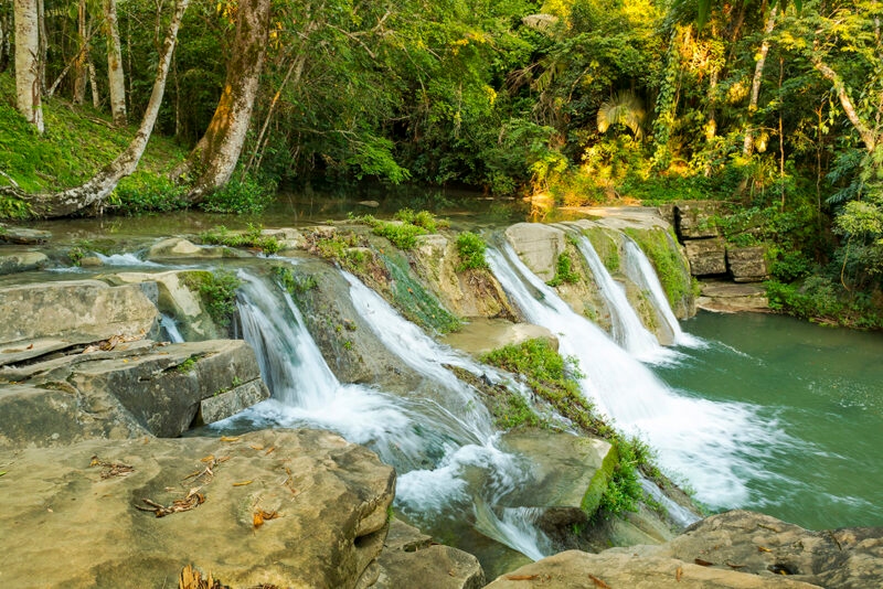 A scenic tropical waterfall flowing into a river within a lush green forest on a sunny day.