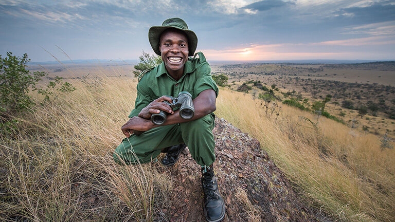 A man crouching in long grass smiling to the camera