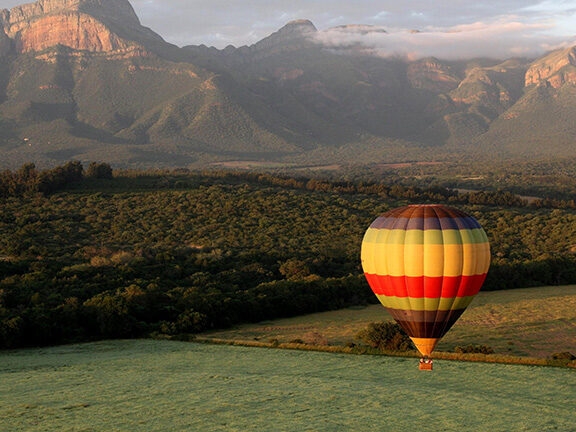 A rainbow striped hot air balloon soaring over fields and forests in South Africa with craggy mountains in the background