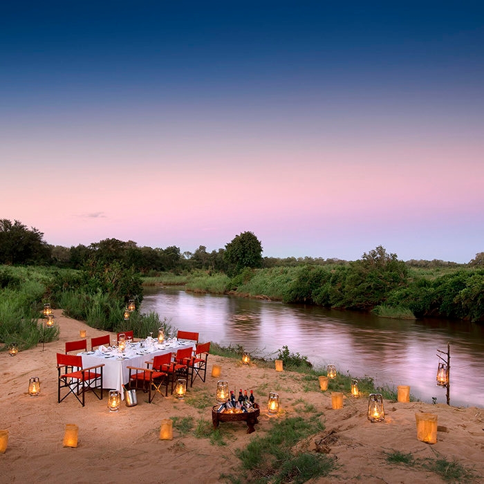 A table set for dinner with chairs and a tablecloth, with lit candles twinkling on the ground, next to a still river