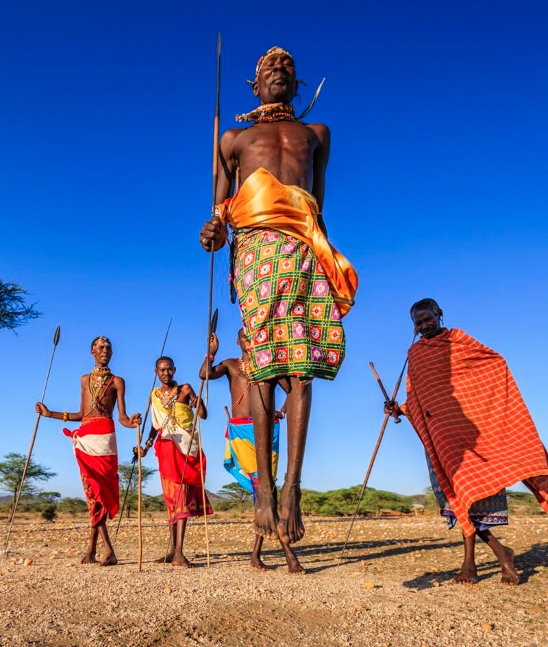 Maasai men in traditional red and patterned clothing performing a jumping dance on luxury Maasai Mara trips.