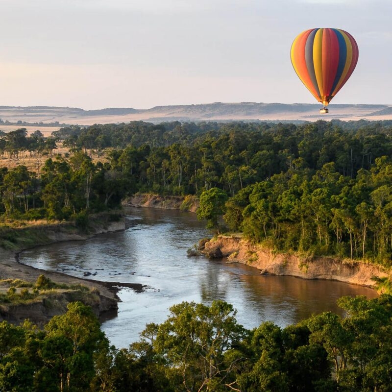 A striped hot air balloon flying over a river and green landscape during luxury Maasai Mara tours.