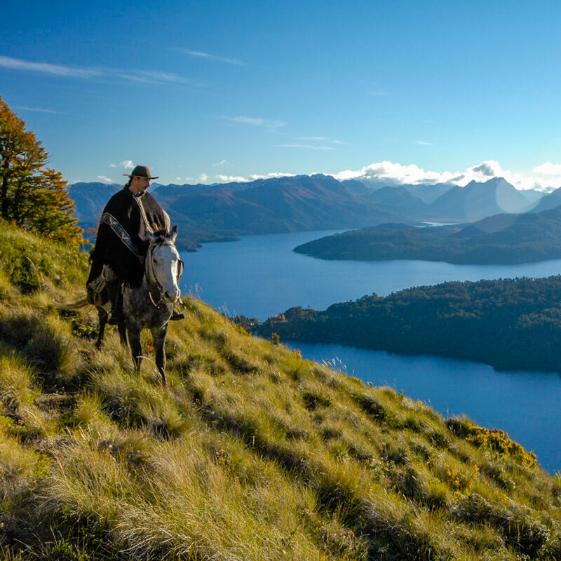 A person riding a horse on a high grassy path above a blue lake