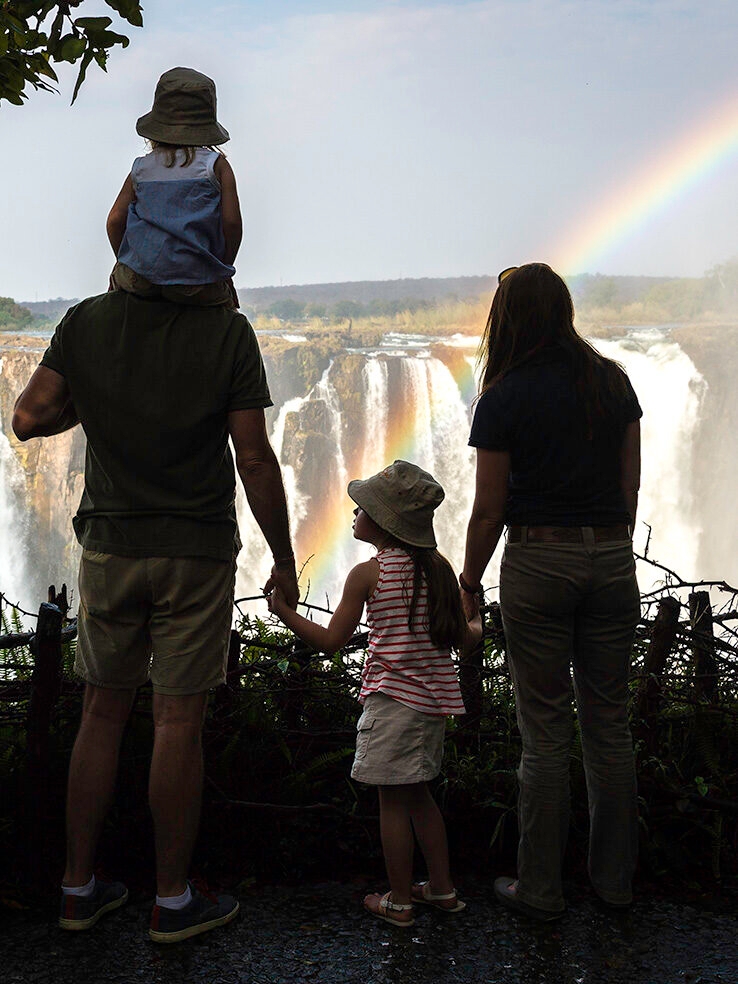 A family standing overlooking the might Victoria Falls in Africa, with a rainbow in front of the water