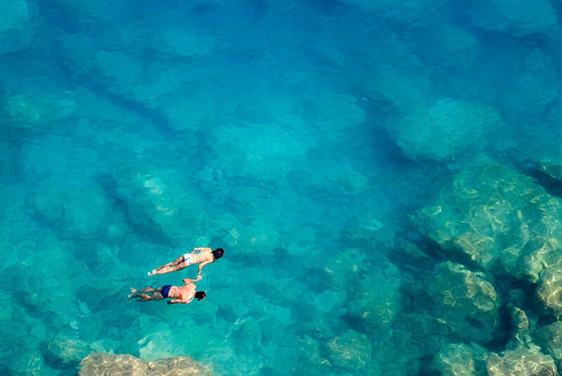 Two snorkellers holding hands while swimming through clear water above a reef