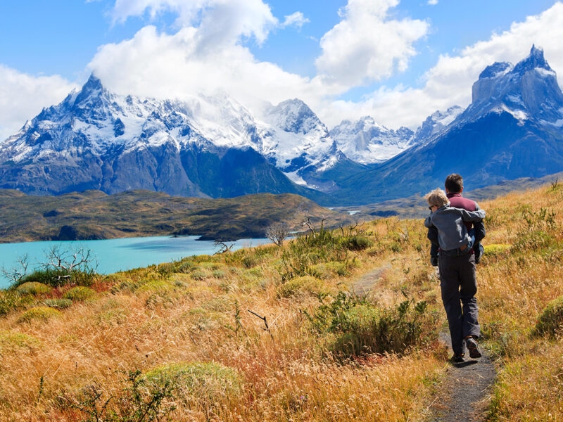 A man carries a child on his back while hiking a mountain trail near a blue lake during luxury Family vacations.